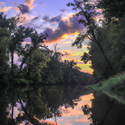 Take a drive along a winding river with a lush forest on one side. Imagine the sun setting, casting a golden hue on the trees and the river's surface. Capture this moment in a romantic landscape painting, with a focus on the vibrant colors of the sky and the reflection of the trees in the water.